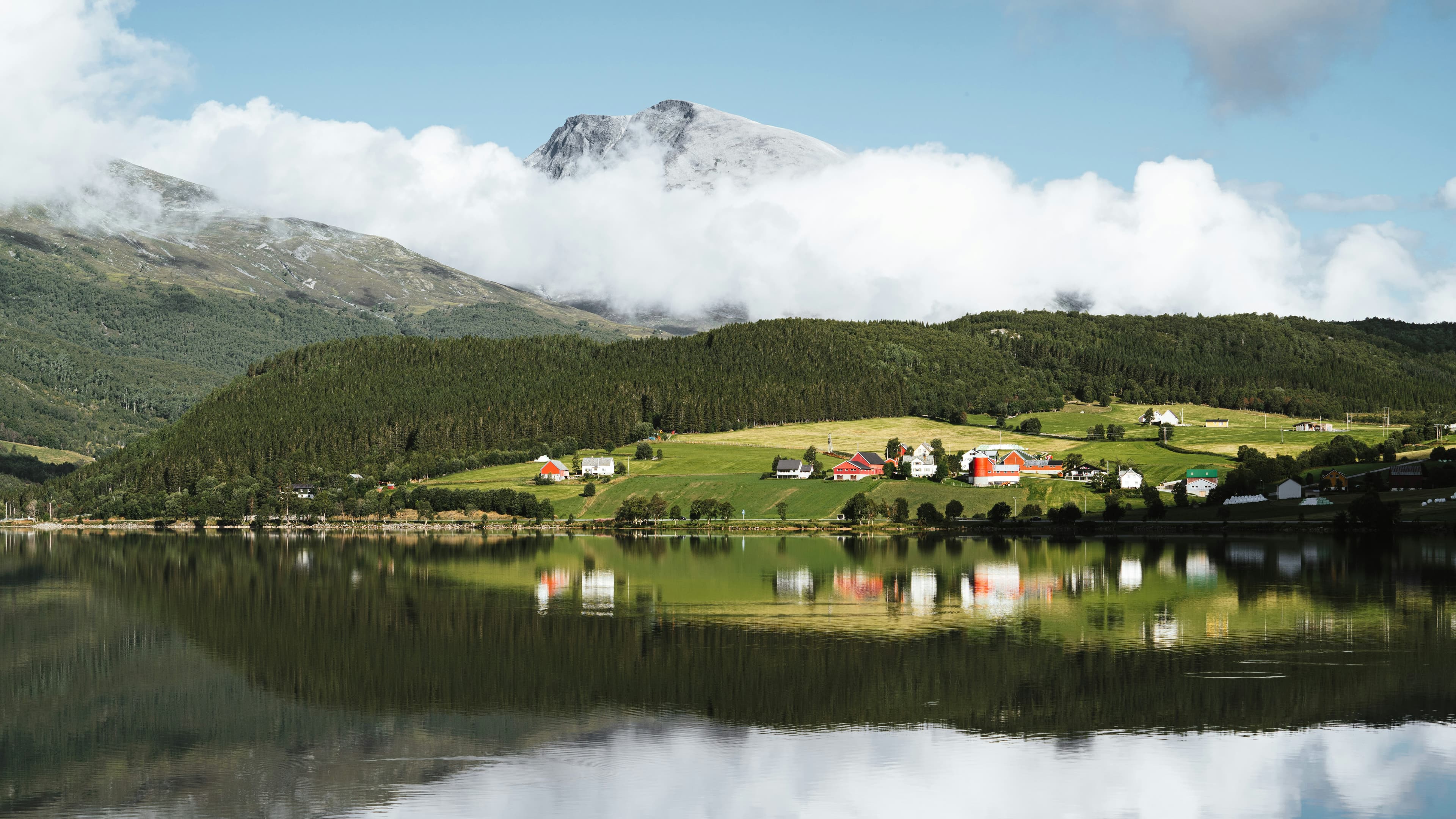 Bygd med røde og hvite hus ved et stille vann, med fjell og en skybanke bak